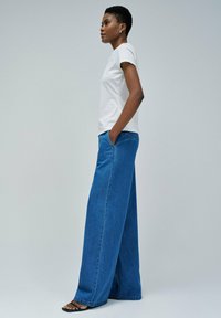 Camiseta de algodón blanca y vaqueros de denim azul de pierna ancha, con bolsillos frontales y un corte holgado, combinados con sandalias planas negras.