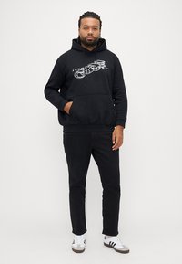 Černá mikina s kapucí s grafickým motivem, přední kapsou a stahovací šňůrkou na kapuci. Kombinováno s černými kalhotami a bílými teniskami s hnědými detaily.