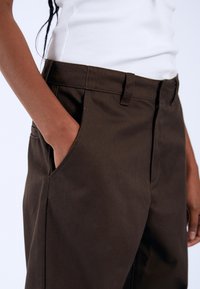 Bruine katoenen broeken met een rechte pasvorm, voorzien van zijzakken en een tailleband zonder zichtbare sluitingen. Glad textuur, minimale details.