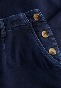 Donkerdenim broek met een close-up van een rits en twee gouden knoopaccenten, voorzien van een gestructureerde stof en een op maat gemaakte tailleband.