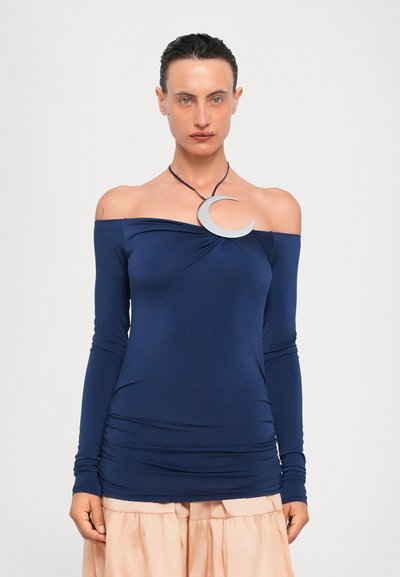 Navyblauwe off-the-shoulder top met lange mouwen en geraffineerde details, gecombineerd met een lichtgewicht roze rok. Zilveren halvemaanvormige ketting die de halslijn accentueert.