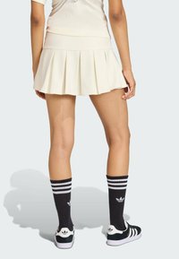 Krémová plisovaná sukňa s rebrovaným pásom, v kombinácii s čiernymi podkolienkami s bielymi pruhmi a logom, a čiernymi teniskami s bielymi akcentmi.