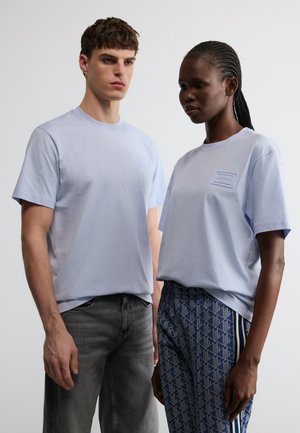 Mladi moški in ženska stojita drug ob drugem, oba nosita svetlomodre srajce; moški v temnih kavbojkah, ženska v vzorčastih modrih hlačah.