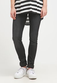 Calças skinny pretas com uma textura suave, combinadas com uma blusa listrada em preto e branco e ténis brancos. As calças têm as bainhas dobradas.