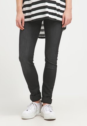Calças skinny pretas com uma textura suave, combinadas com uma blusa listrada em preto e branco e ténis brancos. As calças têm as bainhas dobradas.