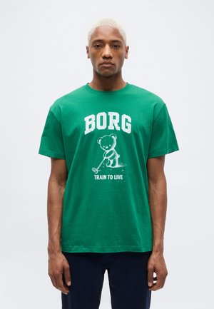 Jeune homme aux cheveux blonds courts portant un t-shirt vert avec un ours jouant au golf entouré des mots « BORG » et « TRAIN TO LIVE ».