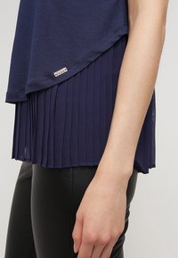 Haut plissé en couches bleu marine avec petite étiquette en métal de la marque, porté avec un pantalon en cuir noir, montrant le bras et la main d'une personne sur le côté.