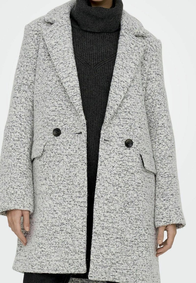 Osoba v šedém vzorovaném dvouřadém kabátu přes tmavě šedý žebrovaný rolák, s viditelnýma rukama po stranách.