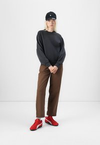 Hanorac negru, pantaloni maro și încălțăminte sport roșie. Căciula este neagră cu un logo alb. Modelul stă cu mâinile încrucișate, fundal neutru.