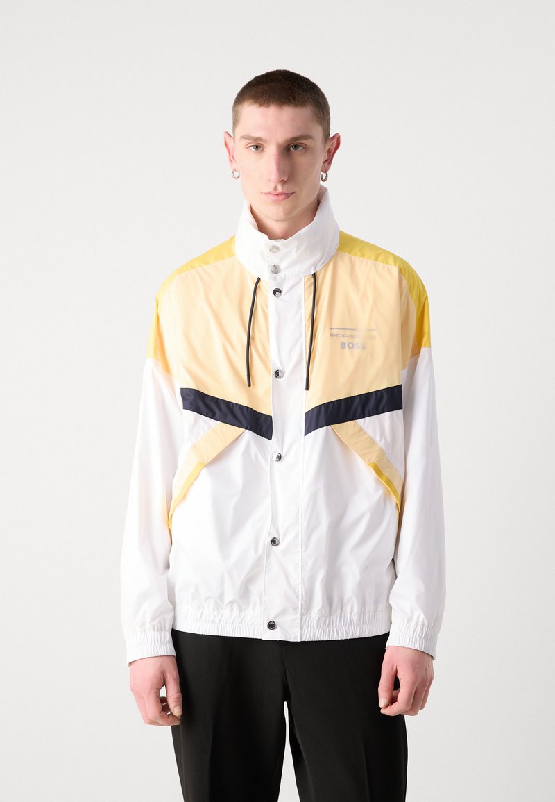 Veste légère blanche avec des accents jaunes et marins, dotée d'un col montant, de boutons sur le devant et de poignets élastiques. Texture lisse, design sportif.