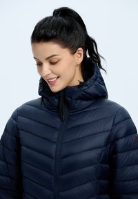 Laivaston sininen puffatakki, jossa on vaakasuora tikkaus, korkea kaulus ja etuvetoketju. Takissa on säädettävät nyörit hupussa ja sileä pinta.