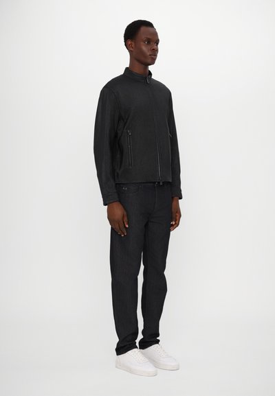 Schwarze strukturierte Jacke mit Stehkragen, mit Reißverschlusstaschen, kombiniert mit dunklen Jeans und weißen Sneakern.