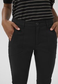 Černé na míru šité kalhoty s hladkou texturou, vybavené dvěma bočními kapsami a zapínáním na jeden knoflík, nošené s černo-bílým pruhovaným topem.
