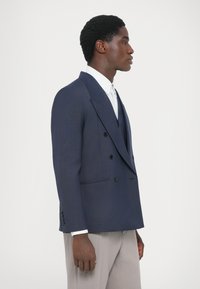 Tengerészeti, dupla soros zakó, lekerekített hajtókákkal, finom textúrával, fekete gombokkal és testhez álló fazonban, fehér ing felett viselve, világos nadrággal.