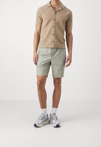 Camicetta a maniche corte color tan con motivo a onde testurizzato, abbottonata, abbinata a pantaloni corti verdi chiari e scarpe sportive grigie con accenti neri.