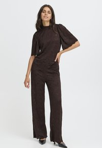 Rudas nėriniuotas kombinezonas su aukštu kaklu, trumpomis pūstomis rankovėmis, plačiomis kojomis ir gėlių raštais. Juodi batai papildo aprangą.