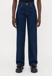 Une personne portant un jean droit bleu foncé et des mocassins noirs épais, se tenant contre un fond clair.