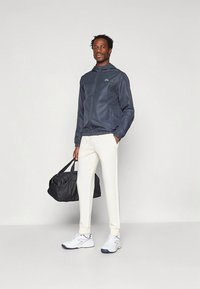 Tamsiai mėlynas vėjo nepraleidžiantis švarkas su gaubtu, baltos kelnės ir juoda sportinė rankinė. Baltos sportbačiai su mėlynais akcentais. Lygus audinys ir prigludęs dizainas.