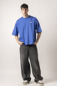 Relaxed-fit blue cotton t-shirt, short sleeves, round neck, with white logo. Paired with loose-fitting gray jeans and sneakers.