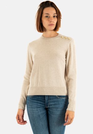 Vrouw met kort bruin haar, gekleed in een beige trui met drie gouden knopen op de schouder en blauwe jeans, staand tegen een witte achtergrond.