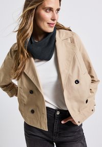 Giacca beige corta con bottoni neri doppi, abbinata a un gilet grigio con collo a cowl e jeans neri. Tessuto liscio, design su misura.