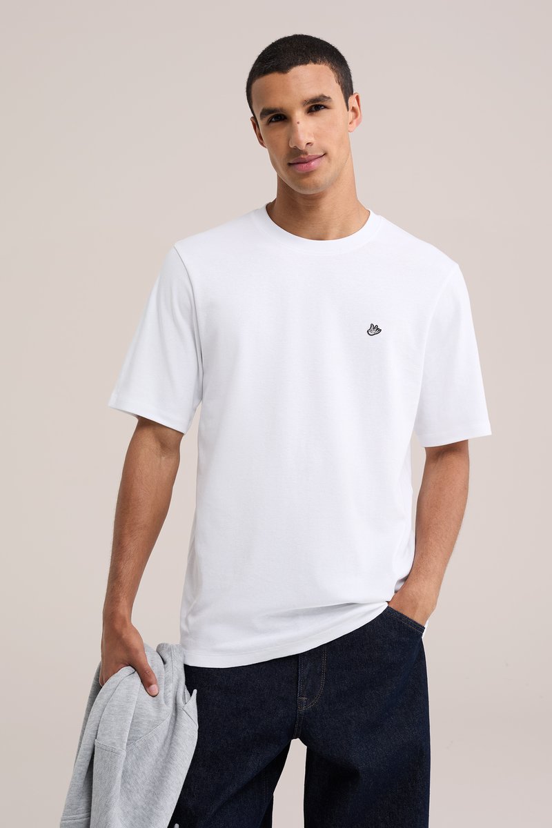 Ung mand iført en ensfarvet hvid t-shirt med lille sort fuglelogo, holder en grå sweatshirt i venstre hånd, stående mod en neutral baggrund.