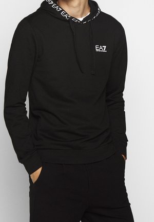 Homme portant un sweat à capuche noir EA7 Emporio Armani avec logo sur la poitrine et capuche, mains dans les poches, sur un fond clair uni.