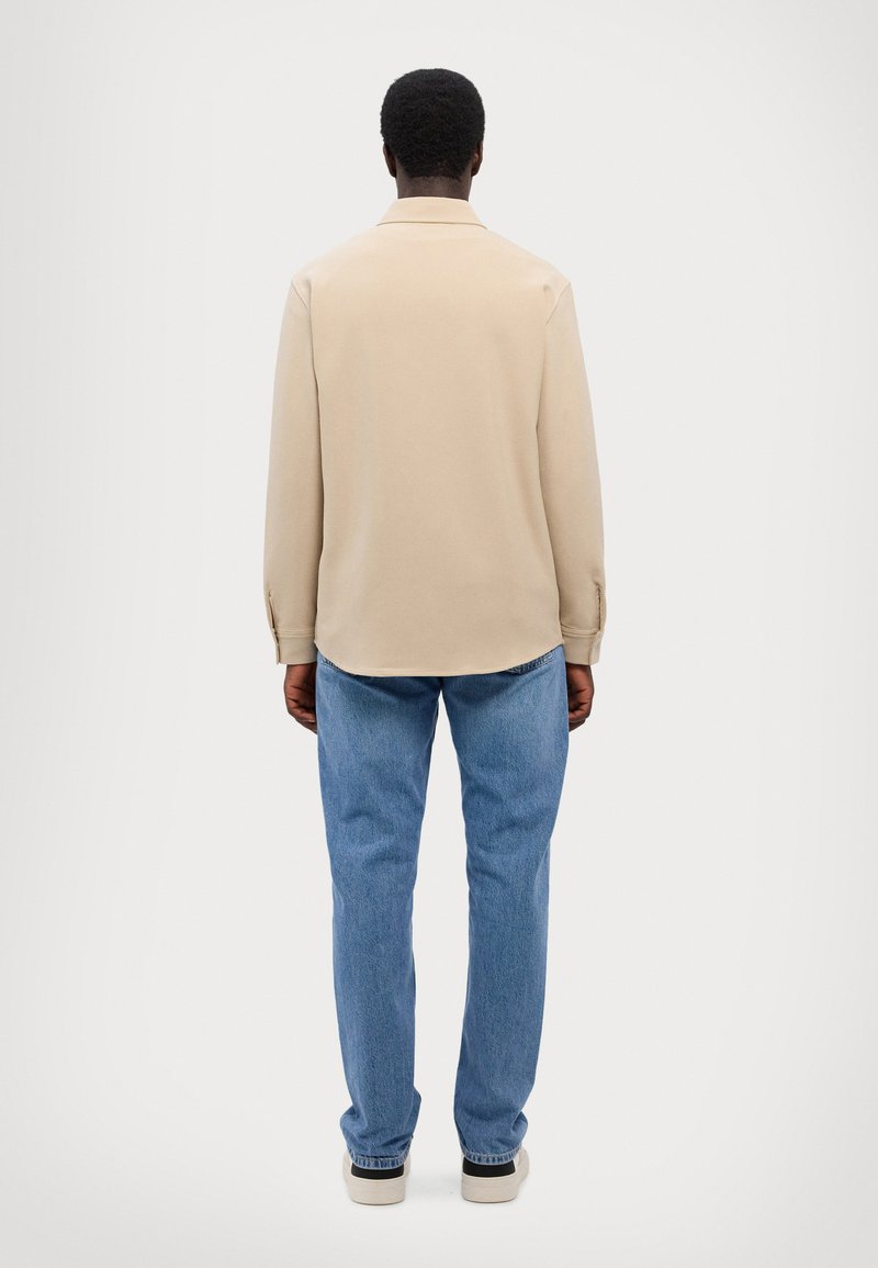 Beige overhemd met lange mouwen en kraag, ontspannen pasvorm en met knopen bij de manchetten, gecombineerd met lichtblauwe jeans en schoenen met zwarte accenten.