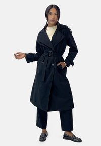 Abrigo negro de trench con grandes solapas, cintura ajustada con cinturón y detalles de botones. Combinado con un suéter crema y pantalones negros, junto con zapatos decorativos.