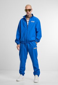 Blue tracksuit made of lightweight fabric, featuring a zippered jacket and elasticized pants. Notable logo patch on the left chest. White sneakers.