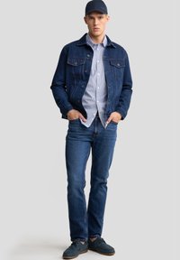 Giacca di jeans, blu scuro, con due tasche sul petto; camicia a righe blu chiaro sotto. I jeans blu slim-fit e le scarpe scure completano il look.