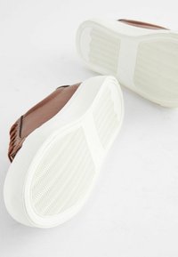 Brune syntetiske sko med en tekstureret overdel, hvide gummisåler og diagonale riller til greb. Designet har en afslappet, slip-on stil.