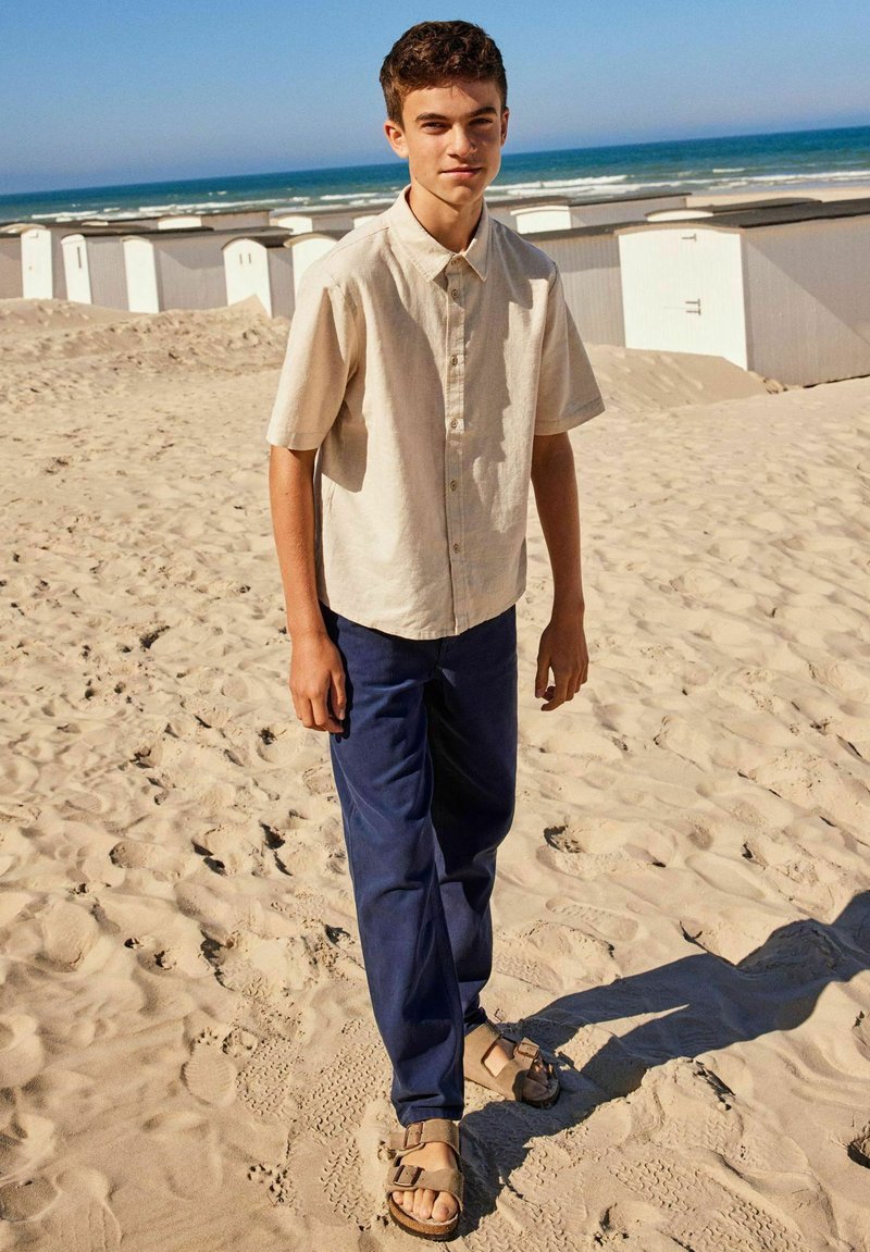 Chemise beige clair à manches courtes, pantalon bleu marine et sandales marron. L'arrière-plan montre une plage de sable avec des cabanes de plage blanches et l'océan.