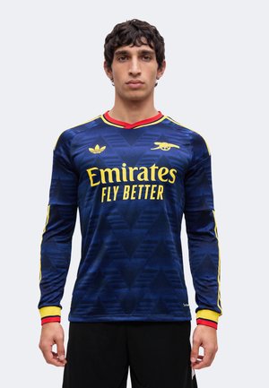 Homme portant un maillot de football Arsenal bleu marine à manches longues avec des bordures jaunes et rouges et le logo "Emirates Fly Better" sur le devant.