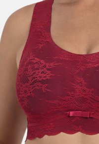 Czerwony koronkowy biustonosz w kwiatowy wzór, z teksturowanej siateczki, szerokimi ramiączkami i satynową kokardą przy podstawie. Wykończenie w postaci falbany dopełnia projekt.