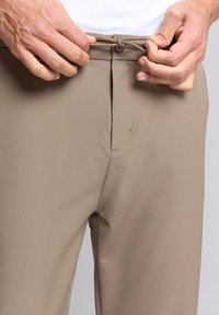 Beige broek met een gladde textuur, voorzien van een knoopsluiting en een trekkoord in de taille. Het ontwerp omvat voorzakken en een getailleerde pasvorm.