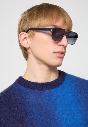 Giovane con capelli castano chiaro che indossa occhiali da sole quadrati scuri e un maglione blu peloso, guardando di lato contro uno sfondo semplice.