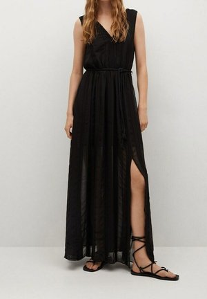Femme portant une robe maxi noire sans manches avec une fente latérale et une ceinture en corde, associée à des sandales plates noires à lacets.