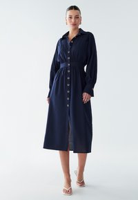 Abito blu navy con maniche lunghe, chiusura frontale con bottoni e vita con cintura. Presenta una gonna con spacco e una texture morbida e liscia.