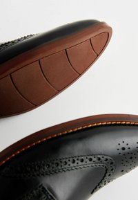 Zapatos de cuero negro con perforaciones decorativas, costura detallada y una suela de goma marrón con patrones de agarre texturizados.