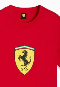 Rotes T-Shirt mit einem gelben Schild-Logo, das ein schwarzes aufsteigendes Pferd und die Buchstaben "S" und "F" darunter zeigt.