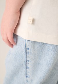 Kremkrāsas kokvilnas t-krekls ar "Mazā putniņa" etiķeti, kopā ar gaiši ziliem džinsa biksēm ar izteiktu sānu šuvi un mīkstu tekstūru.