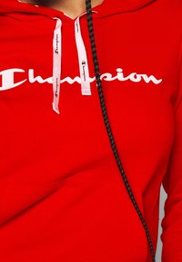 Czerwona bluza z kapturem z miękkiego materiału, z dużym białym logiem "Champion", białymi sznurkami do ściągania z nadrukowaną nazwą marki oraz kieszenią z przodu.