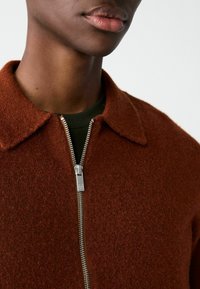 Brown wool blend jacket with a collared neckline, front zip closure, and textured finish. Underneath, a dark green shirt is visible.