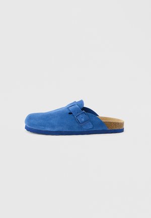 Blauer Wildleder-Slipper-Clog mit verstellbarem Riemen und Schnalle, Kork-Fußbett und profilierter Sohle, von der Seite vor einfarbigem Hintergrund gezeigt.