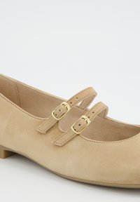 Chaussure Mary Jane en daim beige avec un petit talon, équipée de deux fines sangles maintenues par de petites boucles dorées et un design à bout arrondi.