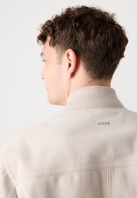 Beež jakk pehmea tekstuuriga, kõrge kraega ja diskreetse logoga taga. Omab minimalistlikku disaini ja õmbluste detaile.