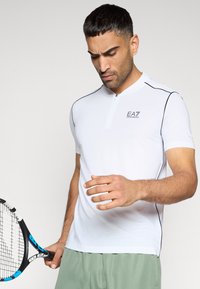 Λευκό πόλο με κοντά μανίκια, ελαφρύ γιακά και φερμουάρ. Διαθέτει μαύρες διακοσμητικές γραμμές και λογότυπο EA7. Το μοντέλο κρατά ρακέτα τένις.