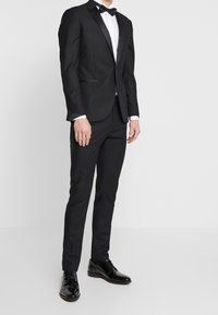 Svart tuxedojacka med satinrevär, parat med matchande byxor och polerade svarta skor. Vit skjorta och svart fluga ingår.