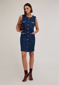 Denim-painettu napillinen mekko, jossa on hihaton muotoilu, etutaskut, kuluneet reunat ja kapea siluetti. Yhdistetty avonaisilla korokepohjilla ja pienellä laukulla.